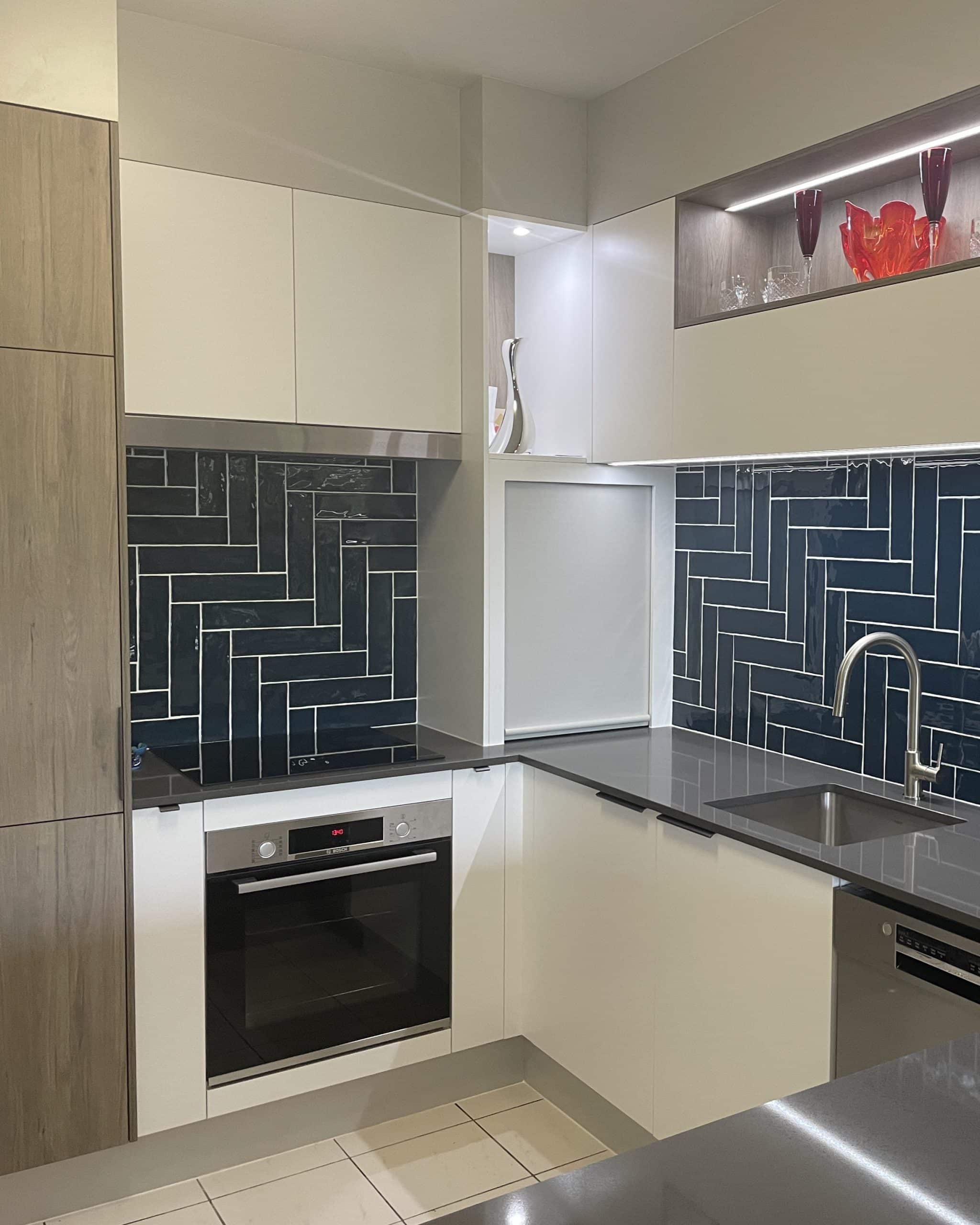 Compact kitchen with turquoise herringbone tile splashback, warm timber finishes and illuminated display shelving — Brisbane kitchen design by Zander Edward